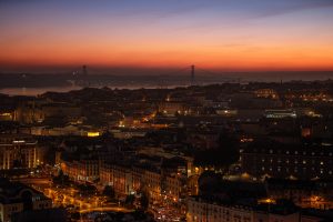 Atardecer sobre Lisboa con edificios iluminados y el Puente 25 de Abril al fondo.