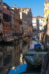 Góndola en canal de Venecia con edificios reflejados en el agua y cielo azul despejado.