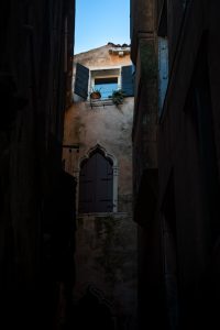 Callejón estrecho con ventana veneciana y contraventanas abiertas iluminadas por el cielo azul.