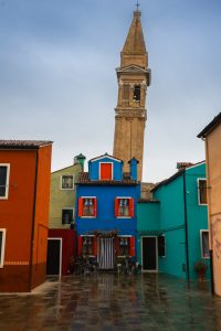Fachada azul vibrante con postigos rojos y la torre inclinada de Burano al fondo, durante una escapada de Murano y Burano en un día.