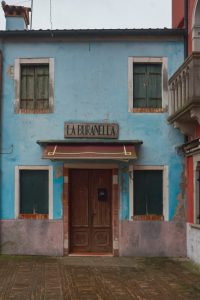 Fachada azul envejecida del local La Buranella en Burano, con ventanas de madera cerradas, capturada en una excursión de Murano y Burano en un día.