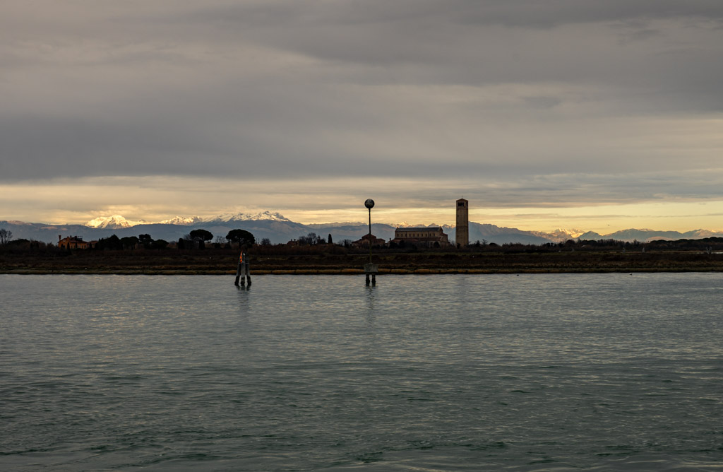 Los Alpes desde la laguna veneciana – Murano y Burano en un díaveneciana-alpes-murano-burano-en-un-dia.jpg Vista panorámica desde la laguna de Venecia con los Alpes al fondo y una iglesia en primer plano, tomada durante una ruta entre Murano y Burano.