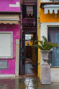 Pasillo entre casas de colores intensos en Burano, con rejas de hierro y un patio al fondo, durante una excursión a Murano y Burano en un día.

